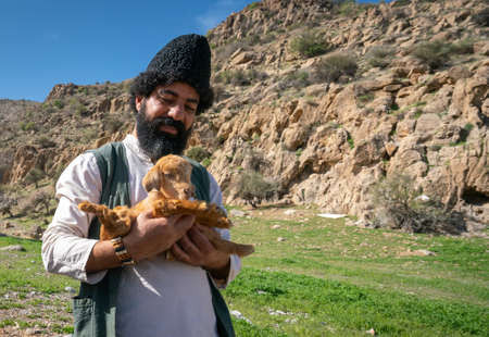 Shiraz, Iran - 04.10.2019: Iranian man with epic beard in traditional hat, white shirt and green vest holds a lamb in his hands. Life of Iranian nomads.のeditorial素材