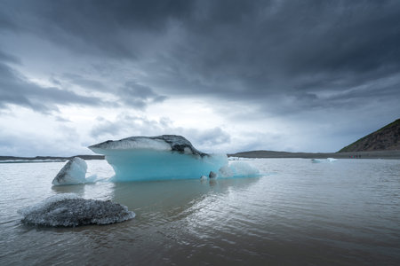 Ice floating in muddy waters of glacier lake with glacier in background on a cloudy and rainy day. Fjallsarlon, Iceland. Tiny people watching from the other side.の写真素材