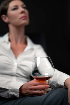 Drinking glass filled with alcohol held by sad looking woman in business clothes sitting with her...