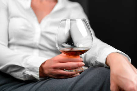 Drinking glass filled with alcohol held by woman in business clothes sitting with her legs crossed. Womans image blurred. Concept for depression, sadness, alcoholism, drinking problems.の写真素材