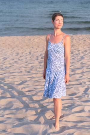 Attractive short haired woman in her thirties wear in light summer dress walks barefoot on the beach with sea in the background. Concept for vacation, leisure, relaxation, physical and mental health.の写真素材