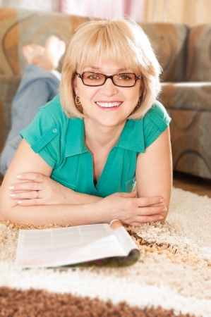 Pretty mature lady lying on carpet smiling  and reading magazineの写真素材