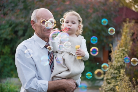 Happy grandfather and cute curly granddaughter blowing soap bubbles