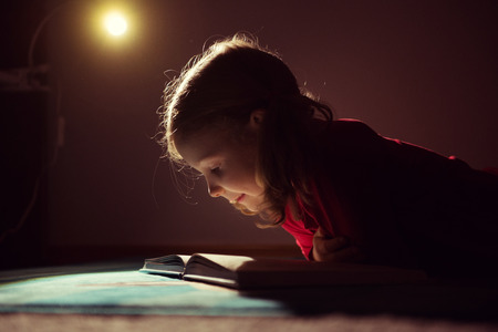 Pretty little girl reading book in her hiding place under bed with flashlight at darknessの写真素材