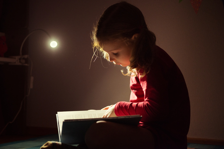 Pretty little girl reading book in her hiding place under bed with flashlight at darknessの写真素材