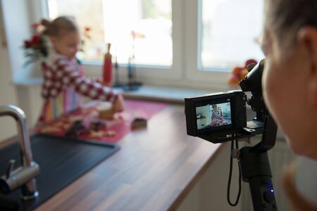 Little pretty girl making christmas ginger cakes in the kitchen and her mother takes photo or video for her blogの写真素材