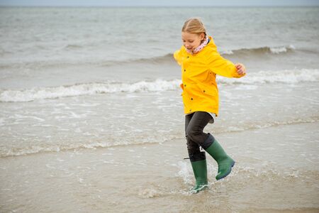 Cheerful little girl running on water of Baltic sea in rubber boots at cold and windy weather 