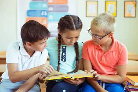 Happy elementary students sitting on desk with book and discussing in classroom at school