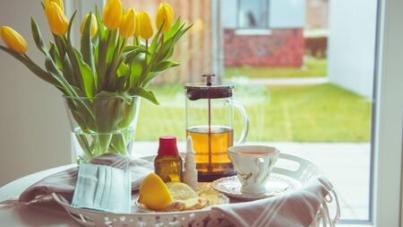 Plate of tea, lemon and ginger on bed table with yellow tulips becouse of corona virus