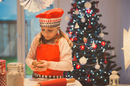 Pretty little girl decoraiting christmas cookies of different forms at home with christmas tree on backgroundの写真素材