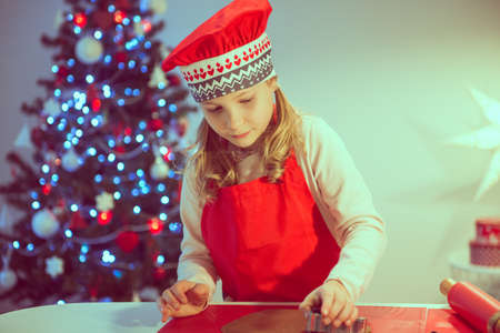 Pretty little girl decoraiting christmas cookies of different forms at home with christmas tree on backgroundの写真素材