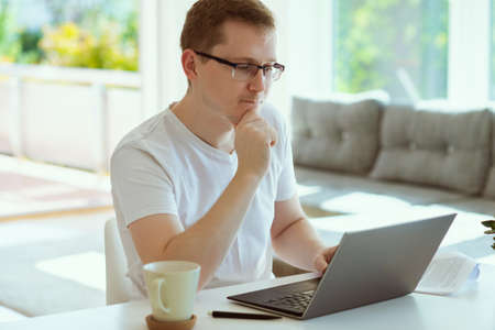 Portrait of serious young man working online with laptop at home, holding paper sitting at office deskの写真素材