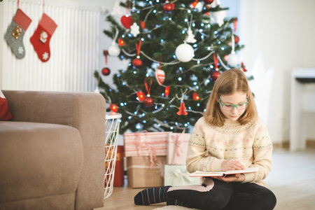 Portrait of pretty little child girl reading book sitting near decorated christmas treeの写真素材