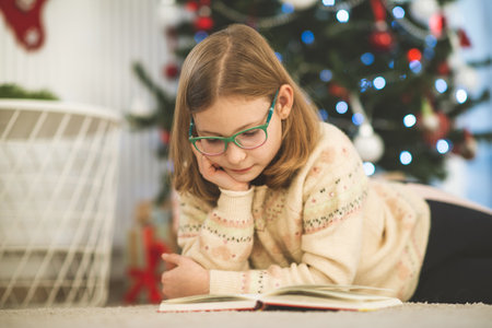 Portrait of pretty little child girl reading book sitting near decorated christmas treeの写真素材