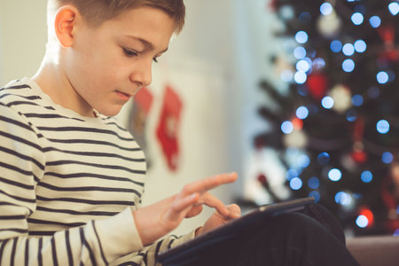 Teenager boy playing or studing with laptop near decorated christmas tree in living room at homeの写真素材