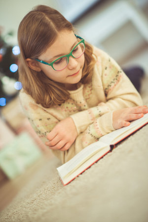 Portrait of pretty little child girl reading book sitting near decorated christmas treeの写真素材