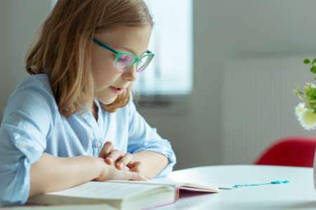 Happy pretty teen girl reading book at desk at home during pandemic distance learning