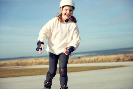 Happy teenager child girl in white helmet, inline skates and safety equipment having fun during learning skating at sunny dayの写真素材