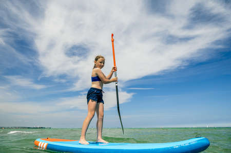 Cute little girl ecercise paddling on SUP board in sea on beautiful sunny summer day while summer holidaysの写真素材