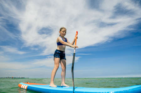 Cute little girl ecercise paddling on SUP board in sea on beautiful sunny summer day while summer holidaysの写真素材