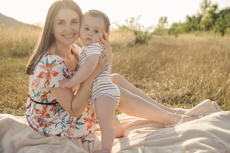 Happy pretty mother with her cute baby son enjoying summer sunlights on blanket in field