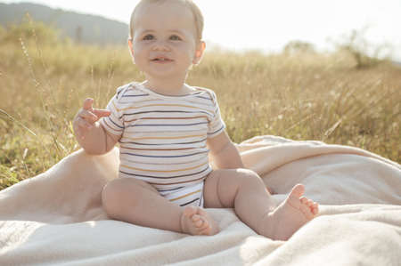 Happy adorable baby boy with beautiful eyes sitting on blanket in field with sunlight on backgroundの写真素材