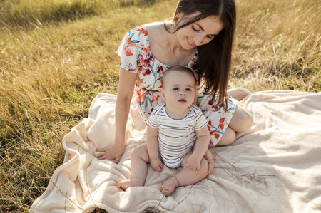 Happy pretty mother with her cute baby son enjoying summer sunlights on blanket in fieldの写真素材