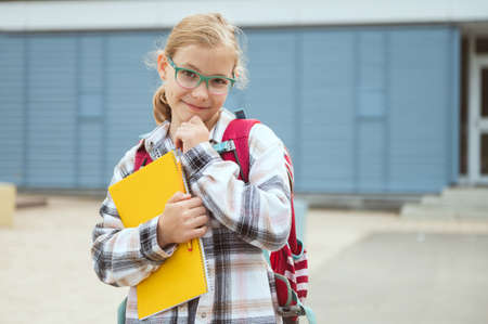 Portrait of pretty little school girl in glasses with backpack standing, posing and smiling in front of schoolの写真素材