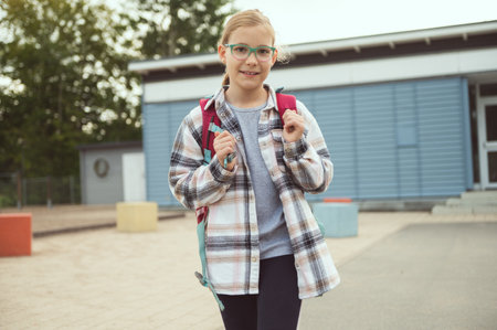Portrait of pretty little school girl in glasses with backpack standing, posing and smiling in front of schoolの写真素材