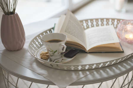 Photo of stylish white tray with black koffee and dark chocoltte and a book. Cozy chair on backgroundの写真素材