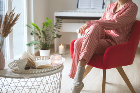 Young woman in elegant pajama sitting on chair near window and tea table with coffee, book and candleの写真素材