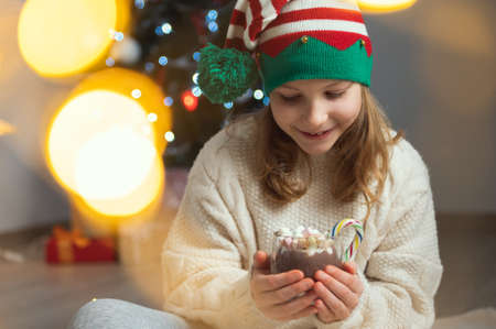 Portrait of happy little child girl holding a cup of hot chocolate with marshmallows with christmas tree and bokeh on backgroundの写真素材