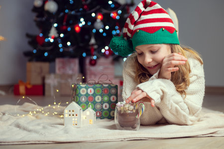 Pretty little girl with funny hat sitting near christmas tree with new years gifts and holding hot chocolate with marshmallowsの写真素材