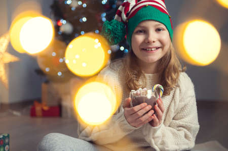 Portrait of happy little child girl holding a cup of hot chocolate with marshmallows with christmas tree and bokeh on backgroundの写真素材