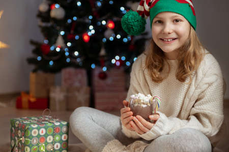 Pretty little girl with funny hat sitting near christmas tree with new years gifts and holding hot chocolate with marshmallowsの写真素材