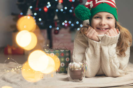 Pretty little girl with funny hat sitting near christmas tree with new years gifts and holding hot chocolate with marshmallowsの写真素材