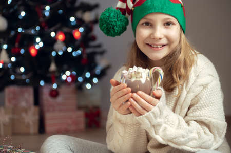 Pretty little girl with funny hat sitting near christmas tree with new years gifts and holding hot chocolate with marshmallowsの写真素材