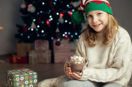 Pretty little girl with funny hat sitting near christmas tree with new years gifts and holding hot chocolate with marshmallowsの写真素材