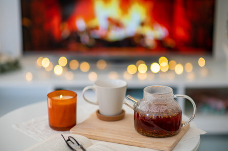 Photo of cozy place with tea cup, teapot, candle and book with glasses with fireplace imitation on...
