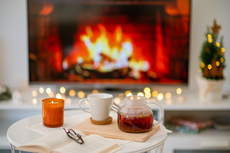 Photo of cozy place with tea cup, teapot, candle and book with glasses with fireplace imitation on tv on backgroundの写真素材