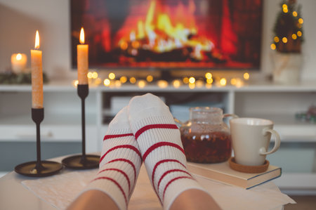 Close up cozy photo of feet in striped socks on side table with candles, teapot and cup  bevor fireplace imitation on TVの写真素材