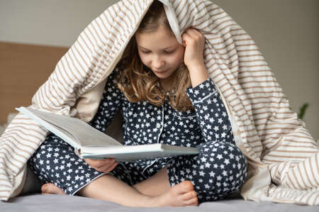 Cute little teen child girl sitting and reading book on bed under the blanketの写真素材