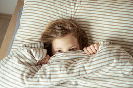 Cute little girl hiding under blanket in cozy bed, just eyes look outの写真素材