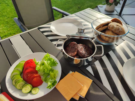 Photo of a table with grilled burgers, vegetables and bread on cozy backyard at summer sunny dayの写真素材