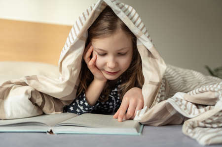 Cute little teen child girl lying and reading book on bed under the blanketの写真素材