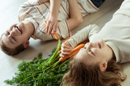 Laughing teen brother and sister lying on the floor with fresh orange carrots in handsの写真素材
