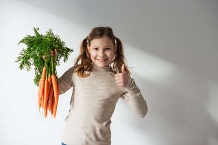 Smiling teen girl holding a bunch of fresh orange carrots with green leaves and showing thumb upの写真素材