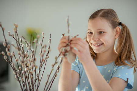 Happy teenage girl hangs easter eggs on tree branches at homeの写真素材