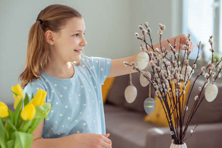 Happy teenage girl hangs easter eggs on tree branches at home
