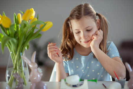 Adorable little girl with two funny pagtails smiling and painting easter eggs at table at homeの写真素材